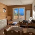 Salon lumineux avec des murs en bois, un canapé, une chaise en osier et une grande porte-fenêtre donnant sur une vue montagneuse.