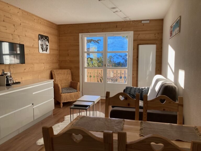 Salon lumineux avec des murs en bois, un canapé, une chaise en osier et une grande porte-fenêtre donnant sur une vue montagneuse.